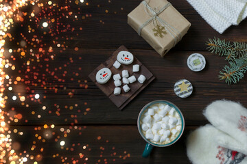 Obraz na płótnie Canvas Christmas New Year card. Cup of hot drink with marshmallows, warm white mittens, New Year's gift, candles on a dark wooden background. Multi-colored bokeh lights. Festive concept. Top view.