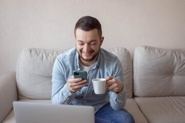 Happy man drinking a cup of coffee at home. Shot of a young man using a smartphone and laptop and having coffee on the sofa at home. Work at home.