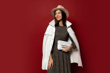 Studio image of gorgeous european brunette gir in trendy white jacket and dress with print posing on red background. Holding leather handbag.
