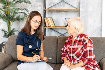 A doctor consulting senior female patient at home. A young female doctor is writing prescription while examining elderly patient