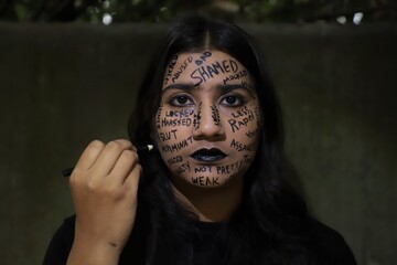 A southeast Asian brown young woman protesting gender based violence by writing anti violence against women and girls messages all over her face