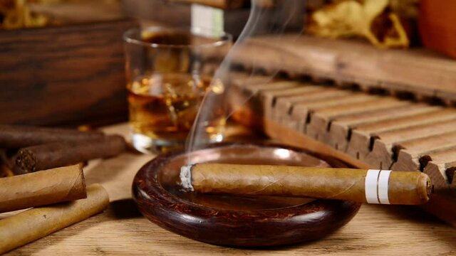 Burning Cigar in an Ashtray with Whisky and Tobacco Leaves on wooden Background