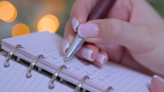 Woman hand putting mark on check boxes of to do list in personal notebook organizer. Garland light bokeh background. Christmas, motivation, management, productivity, planning, reminder, goals concept