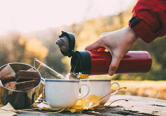 Early morning picnic in forest in late autumn. Cookies and hot tea woman pouring from thermos bottle on wooden table. - obrazy, fototapety, plakaty