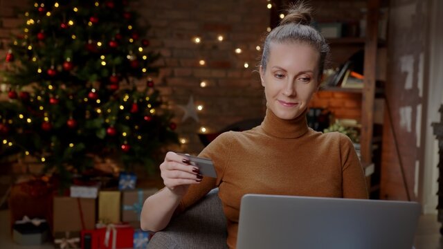 Woman shopping for Christmas gifts online from home in decorated living room with laptop and credit card.