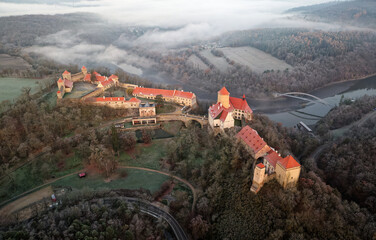 Aerial view of the large and beautiful Moravian royal castle Veveri (Burg Eichhorn), standing on a rock above water dam on the river Svratka, early morning light and autumn weather with the green fore - obrazy, fototapety, plakaty