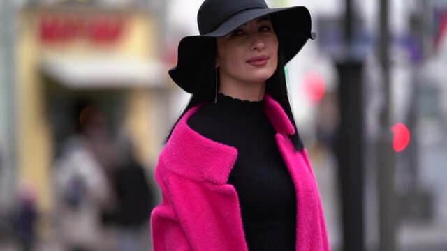 a fashionable woman with black hair, a black hat and a purple coat poses on a street in the city.