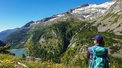 A woman sitting at the side of a mountain and enjoying the view on a lake below at the K?lnbreinsperre in Austria. High peaks are covered with snow. The meadows blossoming with wild flowers. Calmness
