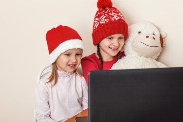 Two little girls in Christmas hats. Two little girls with a soft toy look at the laptop monitor. Two little sisters communicate with relatives online