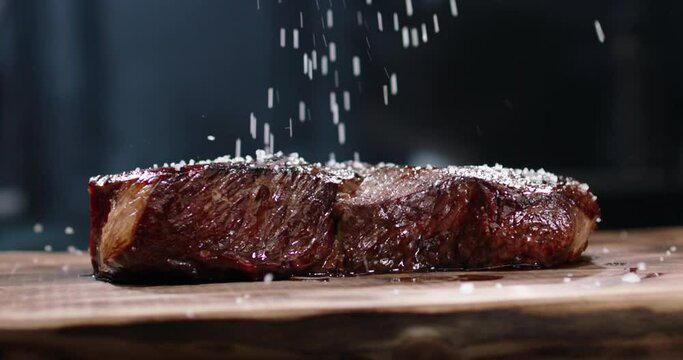 Close up shot of a juicy freshly grilled steak. Big piece of tasty meat straight from the grid. Salt falling onto delicious meal on chef's preparation table 4k footage
