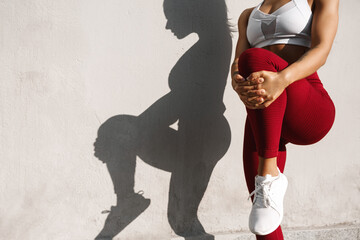 Cropped image of a fitness woman stretching her legs before jogging workout outdoors. Fit female runner doing stretches, warming-up for exercises, shadow on concrete wall - Powered by Adobe