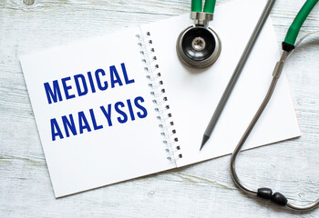 MEDICAL ANALYSIS is written in a notebook on a wooden table next to stethoscope.