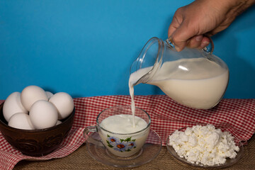 On a blue background, a hand holds a jug of milk. Milk is poured into a cup. Nearby is a plate of cottage cheese and a bowl of eggs.