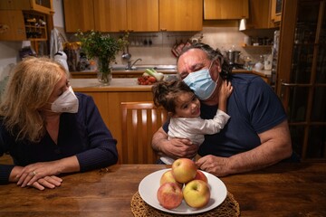granddaughter hugs her grandparents as they sit at the kitchen table wearing health masks