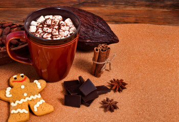 Cup of cacao drink with marshmallow, gingerbread man, chocolate, spices, cocoa pods and cocoa beans on a cork background.