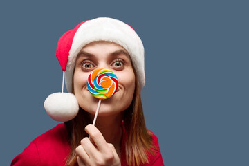 Fototapeta premium Beautiful cheerful girl in a red Santa hat with a lollipop instead of a nose on a blue background, close-up, copy space. Celebrating new year or christmas