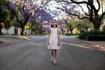 A cute little girl in a white dress walks down the street with blooming jacaranda trees