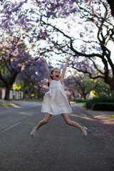 A little cute girl with long hair in a white dress jumps on the street with blooming jacaranda trees