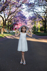 A cute little girl with long hair in a white dress stands outside with blooming jacaranda trees