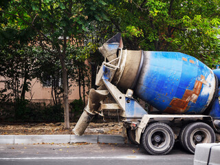 A blue concrete mixer stands on the side of the road near the curb against the backdrop of a house...