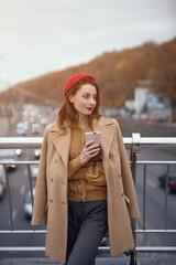 Tender young woman warming up with cup of coffee on the street female fashion standing on a pedestrian bridge. Portrait of stylish young woman wearing autumn coat and red beret outdoors. Toned image. 