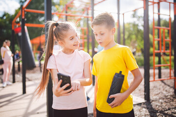Fototapeta na wymiar Caucasian children teenagers use cell phone in workout at street gym. Diverse twins watching video lessons with gymnastics exercises. Brother and sister use exercise app on smartphone by themselves