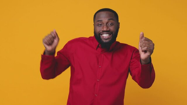 Smiling cheerful excited young bearded african american man 20s wearing red shirt isolated on bright yellow color background studio. People lifestyle concept. Looking camera dancing clenching fists