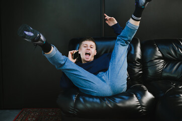 Playful young man in casual clothes is lying upside down on a black leather sofa, talking on the phone, looking into the camera and smiling. Guy is calling on the phone and having fun on the couch.