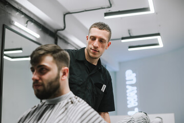 Portrait of a positive male hairdresser in the process, creating a fashionable hairstyle for a bearded adult client. Barber cuts a man with a beard and smiles. Barbershop concept.