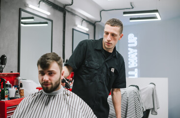 Naklejka na ściany i meble Portrait of professional barber working on haircut of adult man with beard in light barber shop. Serious male hairdresser creates a stylish hairstyle for the client with the help of a clipper.