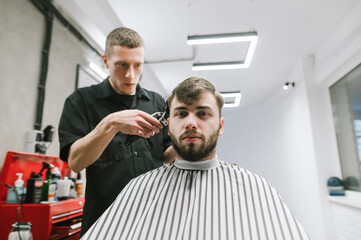 Adult bearded man in a dressing room sits in a barbershop chair, cuts hair at a hairdresser, looks in camera with a serious face.Concentrated barber cuts a brunette client with a clipper in her hand.