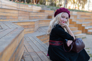 A joyful woman sits on a summer theater bench, made of wood in a burgundy coat and biret, with beautiful eyes, in the fall against the background of trees blue clouds.