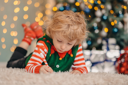 Dreamy little boy writing a letter to Santa Claus lying on the floor in a decorated room, waiting for gifts and a miracle