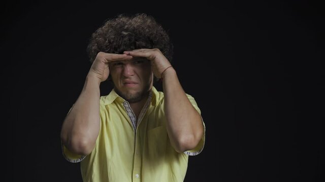Looking for something young guy with long curly hair use hands to cover from lights isolated against a black background. 4K High definition footage. 