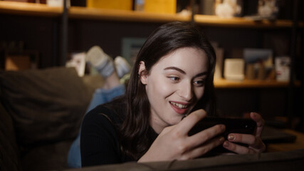 Pretty girl is lying on the sofa and playing games or watching videos with her mobile smart phone in hand, social life and technology concept