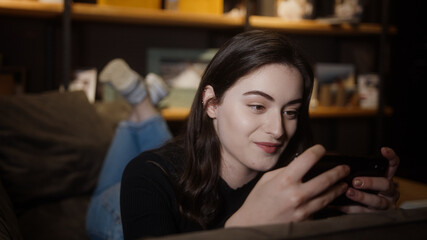 Pretty girl is lying on the sofa and playing games or watching videos with her mobile smart phone in hand, social life and technology concept