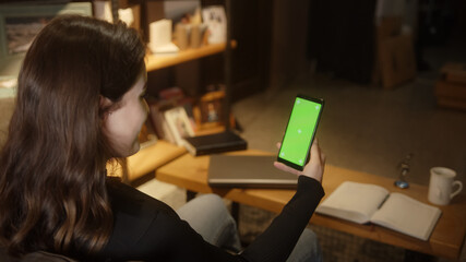 Woman with brown hair is sitting on the sofa at home, holding the green screen phone in her hand and she making video chat, green-screen mockup