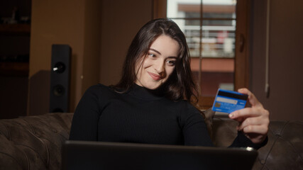 Young beautiful sweet girl sitting on the sofa is shopping on her laptop and credit card in hand, shopping and technology concept