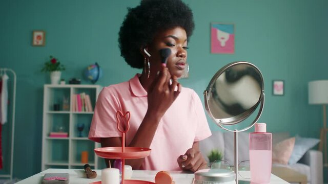 Young black woman is sitting at beauty table at home, getting ready to go outside, doing makeup, uses makeup brush to apply cosmetics, Foreground, Slow motion.