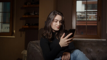 Fototapeta premium Pretty girl sitting on sofa and texting or shopping with mobile smart phone, social life and technology concept