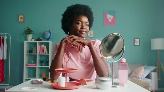 Portrait of young Afro-American woman sitting at beauty table with different cosmetic and care stuff, in pink T-shirt and big earrings, looking on camera and smiling, Slow motion.