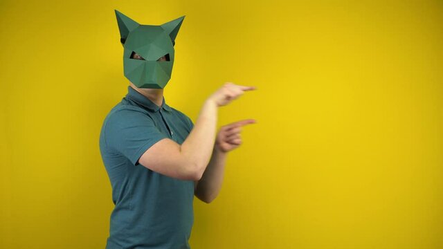 A young man in a cardboard jackal mask points to the empty space with his index fingers on a yellow background. Man in a green polo and mask.