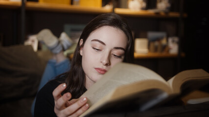 Sweet young woman lying on sofa at home, reading novel or educational book in hand, reading and education concept