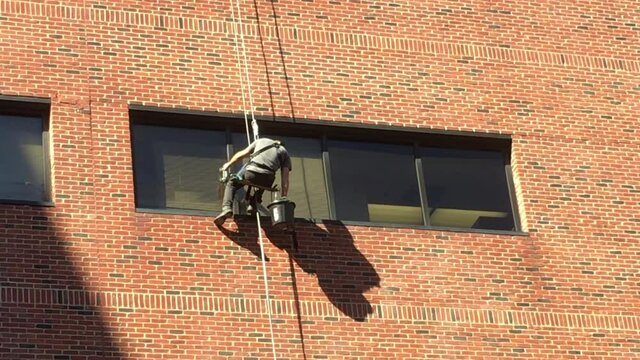 Commercial window washer sits on platform also known as bosun's chair and leans back with right hand to get soapy water on brush, then cleans. His left hand holds a suction positioning handle. 4K