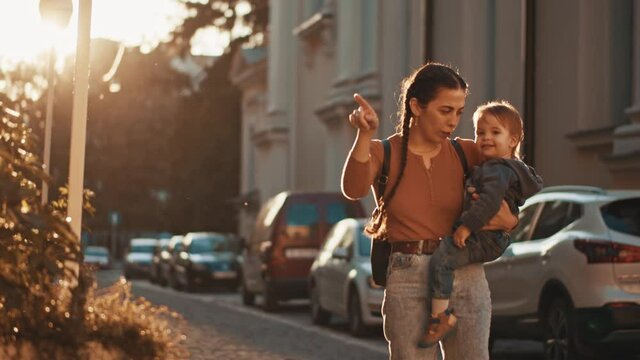 Young and cheerful mom walking along sunny street with small cute baby in her arms. She is happy to be a mother and raise a child. They saw friend or dad and wave to him. Carefree childhood, parenting