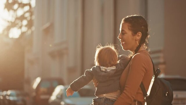 So fun together! Young adorable mother with backpack revolving around her axis with her little cute son while walking at the sunny street and feeling pleasure emotions. Happy childhood, parenthood