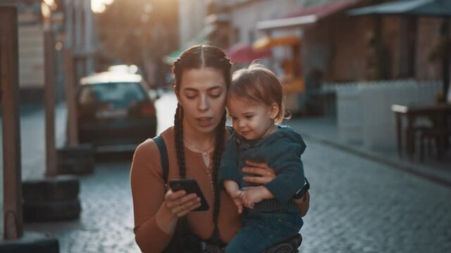 Attractive young mom with pigtails hugging and holding her adorable kid while walking on the street of old city. Use smartphone watch cartoons, funny pictures, play game. Happy parent, mom and child