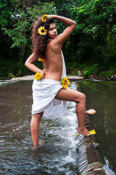 Girl with dark skin and curly hair dressed in a white cloth and a naked torso surrounded by sunflowers and painted face in a river in a forest