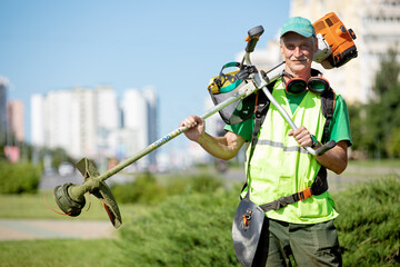 Municipal gardener landscaper senior man worker with gas grass trimmer equipment on sunlight city...