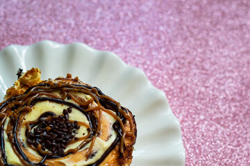 Cinnamon rolls with various sweet toppings and chocolate on a white plate on the pink glitter background.